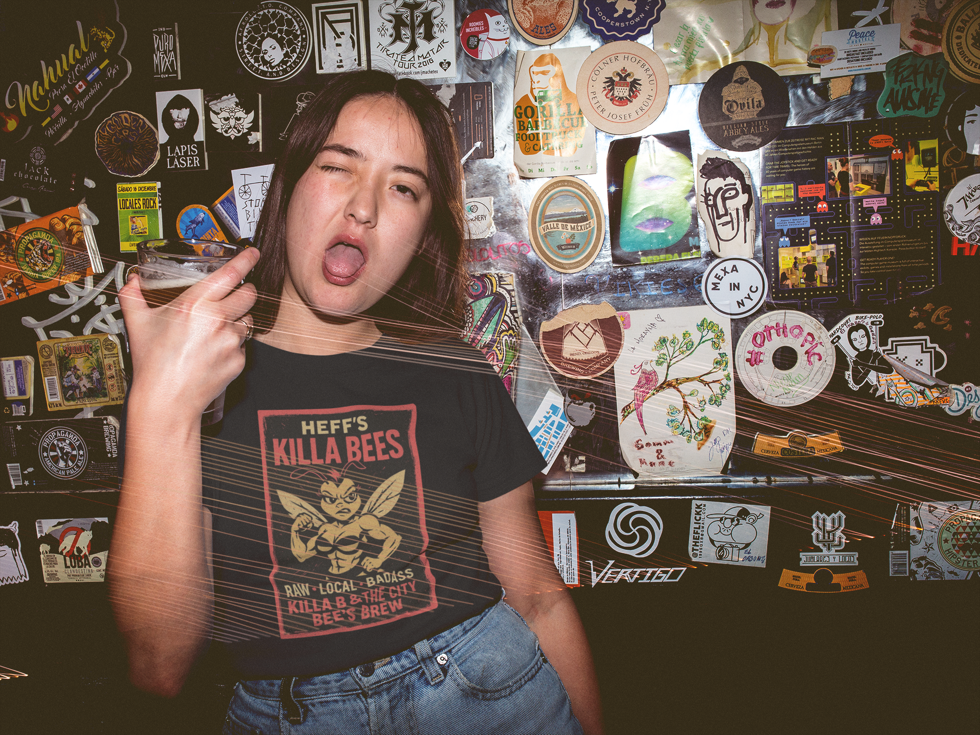Person wearing a black t-shirt with 'Heff's Killa Bees Honey OG' graphic in front of a wall covered in various stickers.