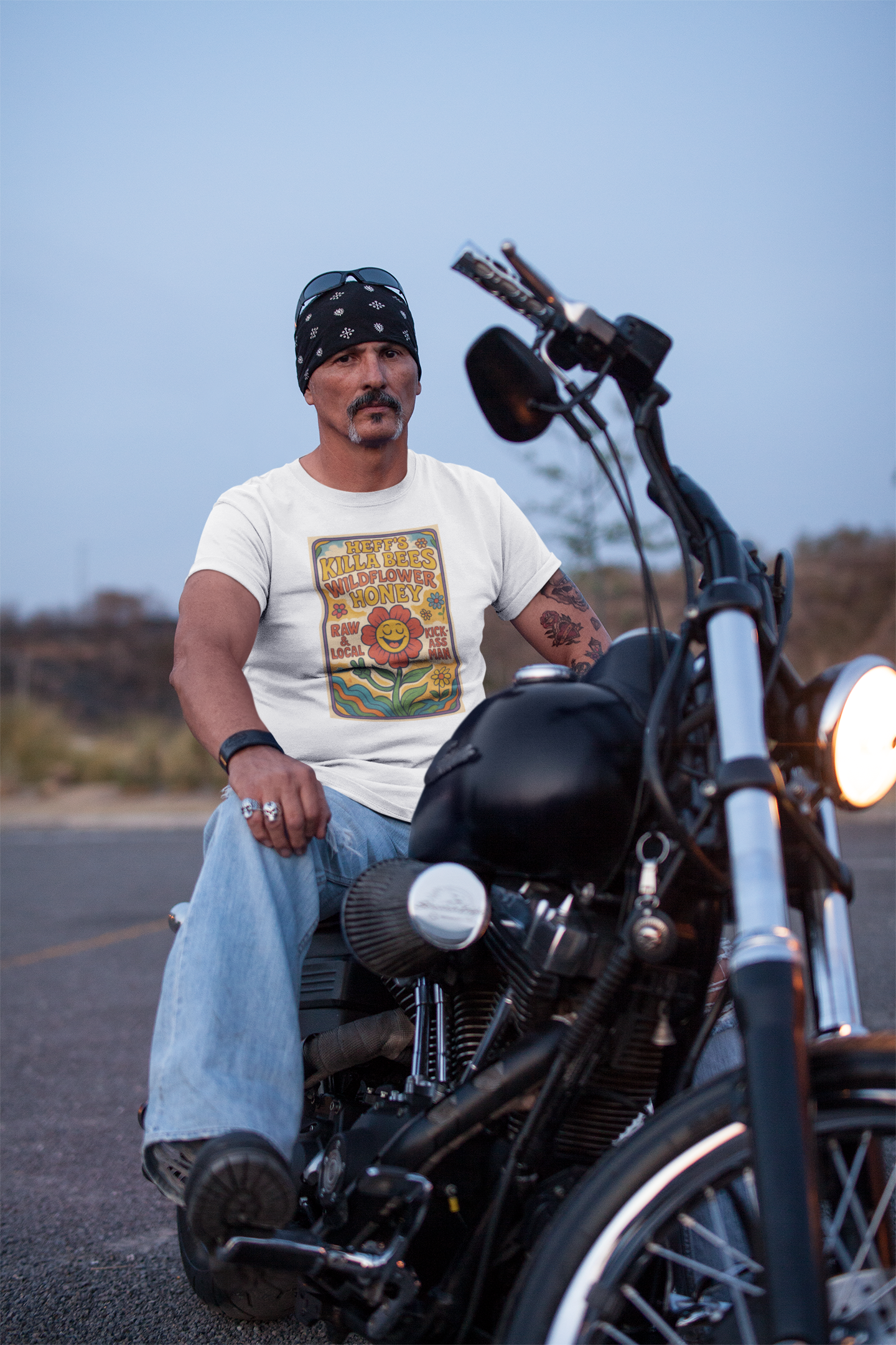 Man sitting on a motorcycle wearing a bandana and a graphic t-shirt, with a clear sky background.